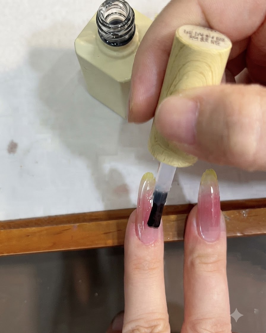 A close-up of a brush applying a clear top coat over a long nail with a pink and yellow gradient.