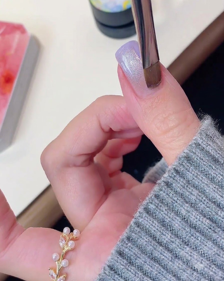 A nail brush or magnetic tool hovering over a purple and shimmering cat-eye gel nail.