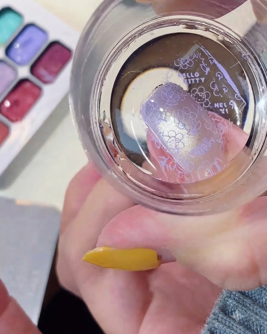 A clear silicone stamper transferring a purple Hello Kitty pattern onto a manicured nail.