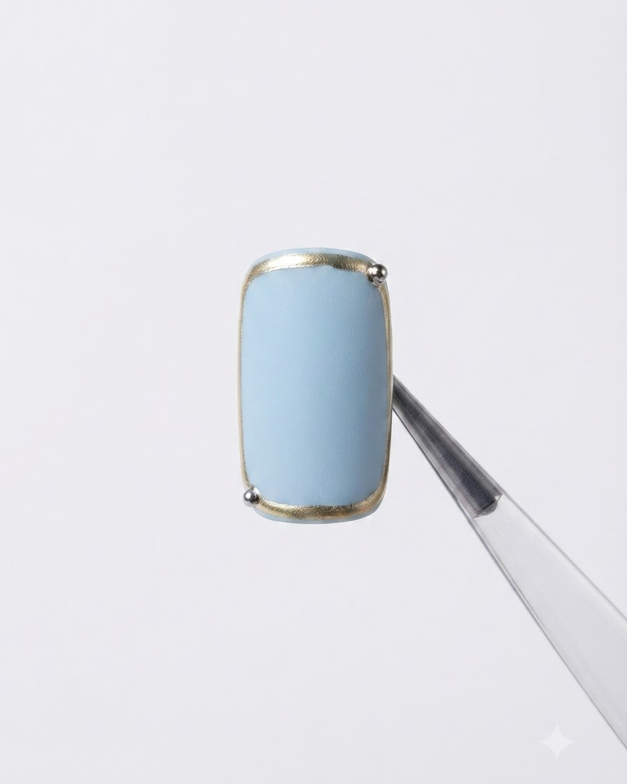 A nail held with tweezers featuring a finished metallic frame border and a tiny silver bead at the corner.