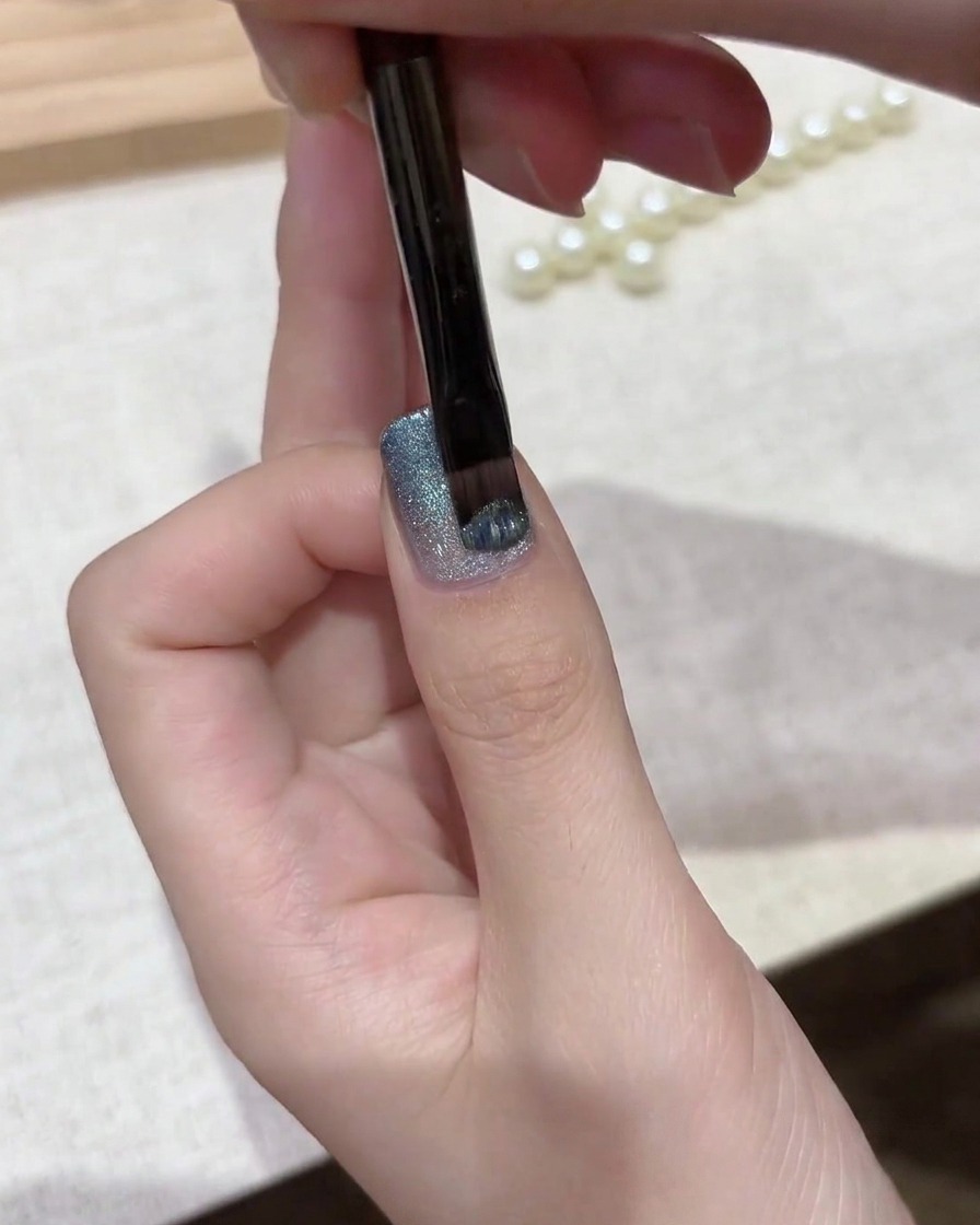 A gel brush applying a coat of shimmering silver-blue magnetic cat eye polish over a gradient base.