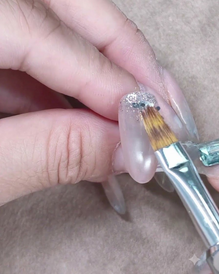 A small nail brush gently feathering silver glitter downwards on a clear gel nail.