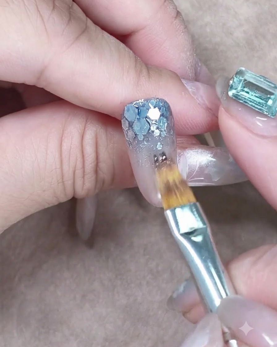 A flat brush dabbing sheer blue gel onto chunky silver sequins at the nail base.