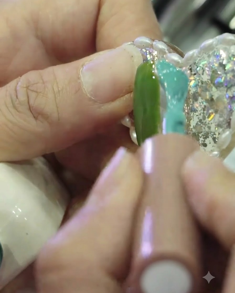 A close-up of a nail brush applying teal gel polish to the right side of a nail, with the left side painted green, leaving a narrow unpainted strip in the center.
