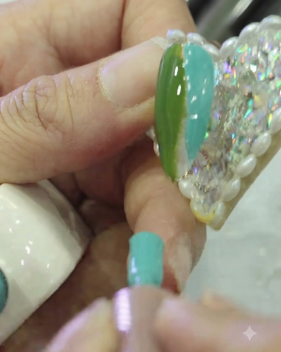 A close-up of a flat nail art brush dabbing the boundary between dark green and teal gel polish on a nail.