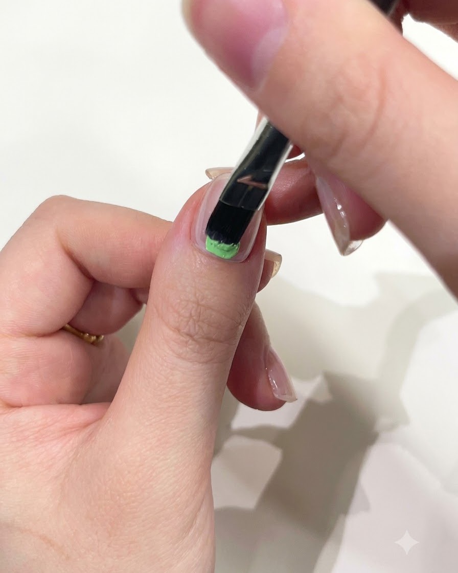 A close-up of a hand using a flat gel brush to apply mint green solid gel to the base of a thumb nail.