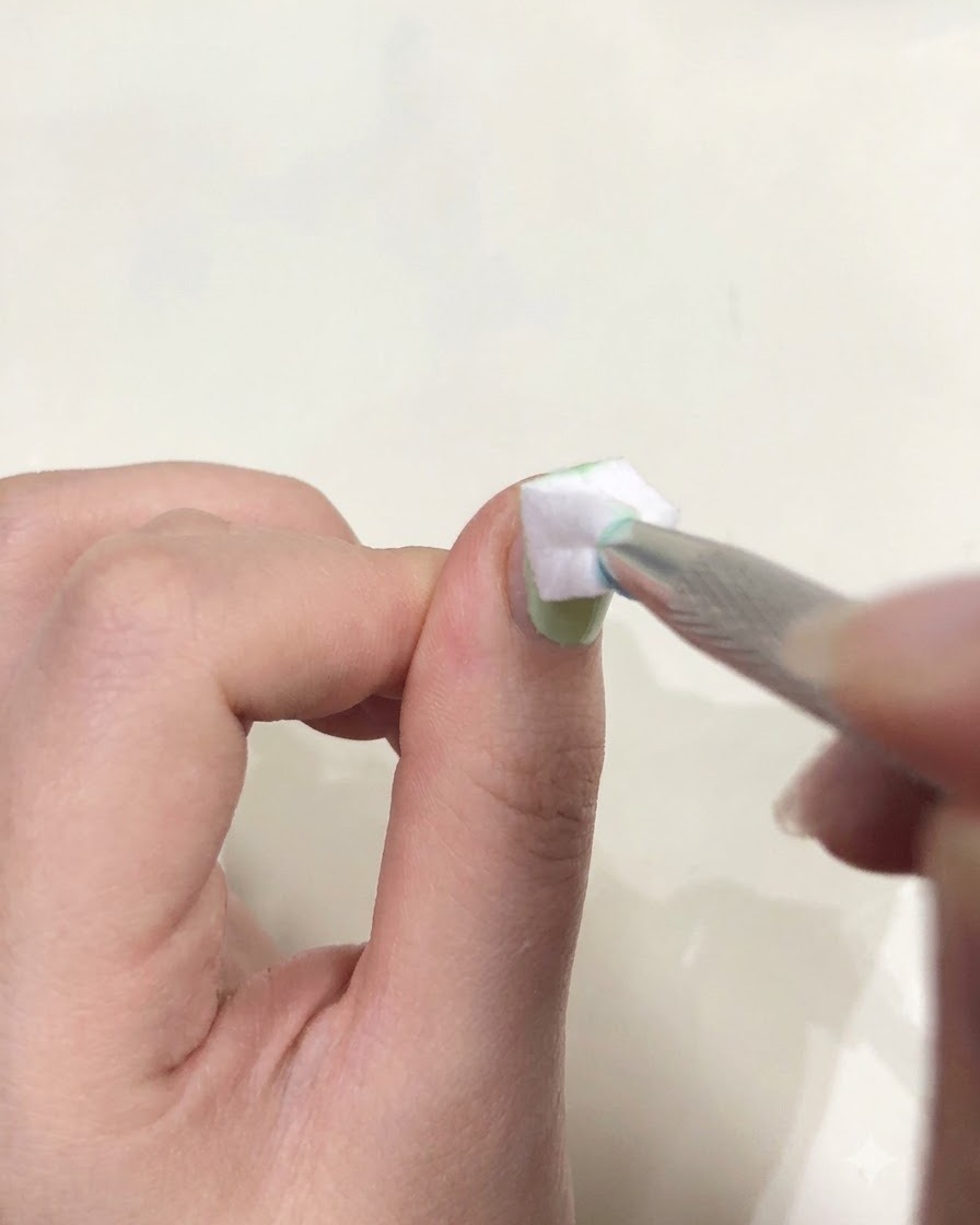 A close-up of a hand holding tweezers with a small white sponge dabbing green gel on a fingernail.