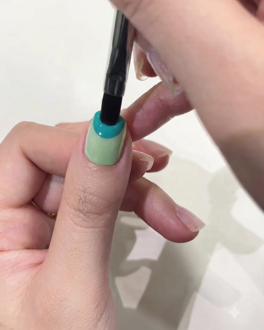 A close-up of a flat gel brush applying pastel blue solid gel to the top half of a thumb nail.