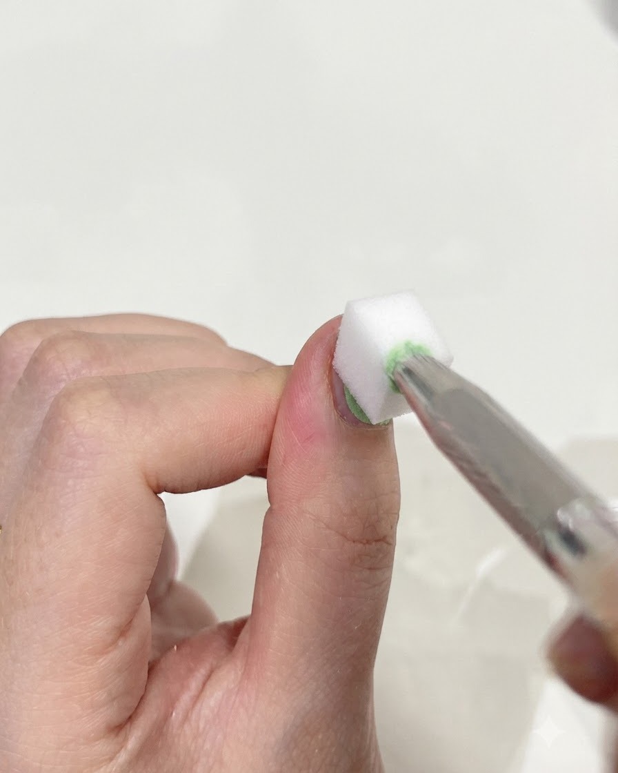 A close-up of tweezers holding a small white makeup sponge, gently dabbing the surface of a thumbnail to blend green and blue gels together.