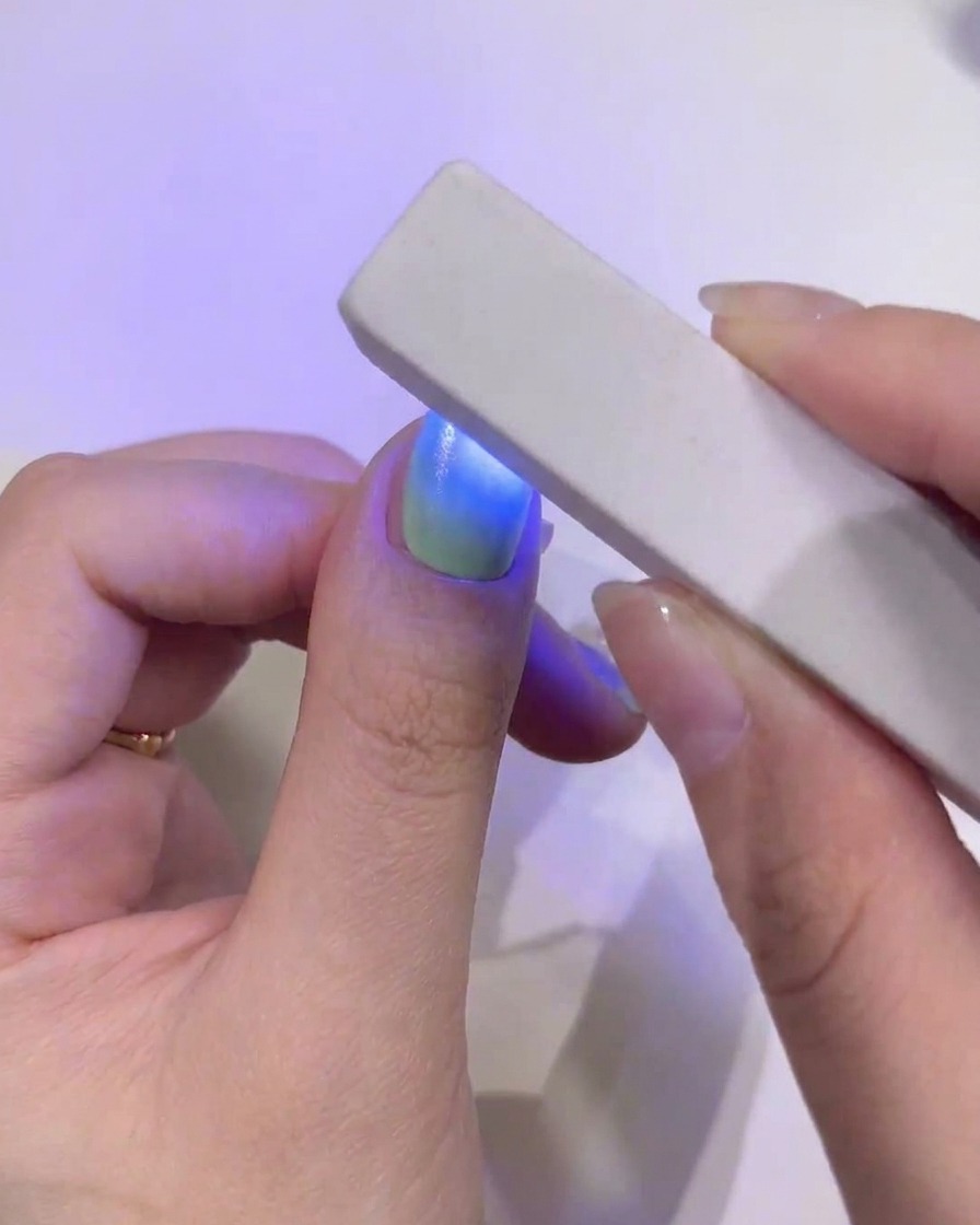 A close-up of a thumb nail with a seamless green and blue gradient being cured under a rectangular mini handheld blue UV/LED lamp.
