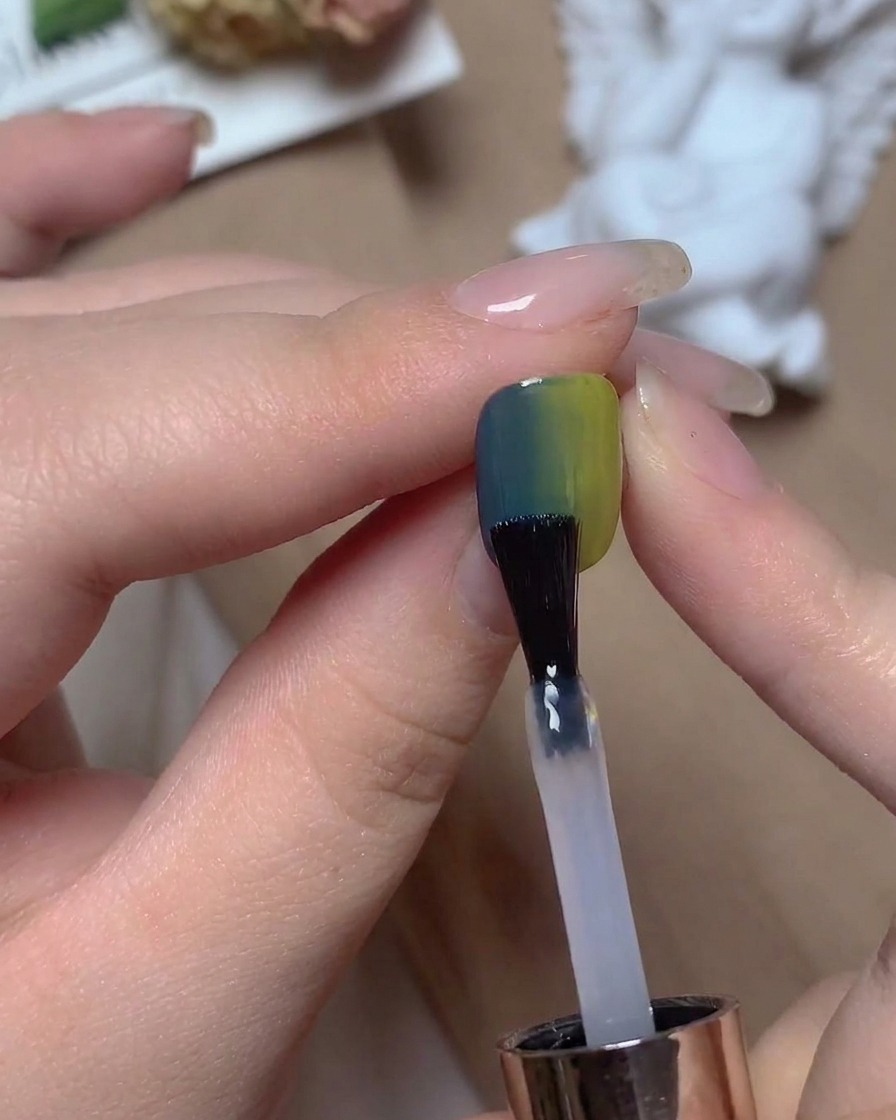 A nail brush applying a clear top coat over a blue and green gradient nail under warm studio lighting.