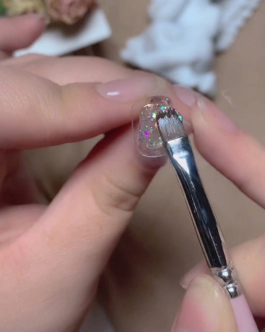 A fine brush applying sparkly multi-colored glitter gel onto a nail surface with vibrant reflections.