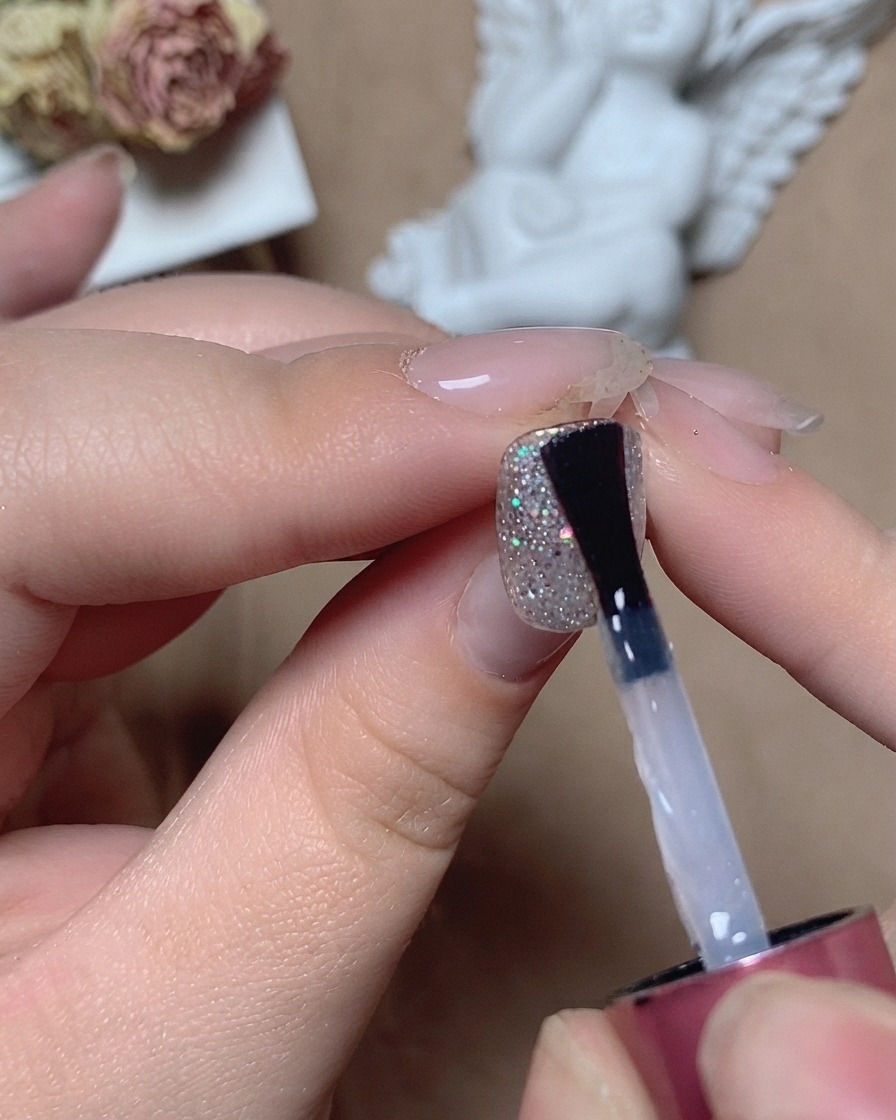 A brush applying a clear leveling top coat over a textured glitter nail under studio lighting.