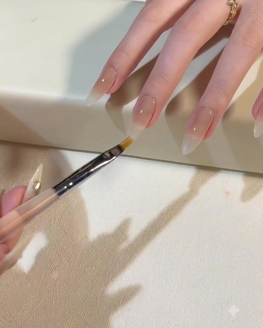 A brush applying milky white gel to create an ombre gradient fading down the nail.