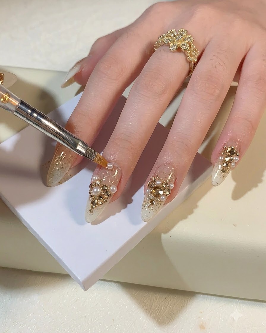A detail brush or tool placing small pearls and champagne gold rhinestones onto a glitter nail.