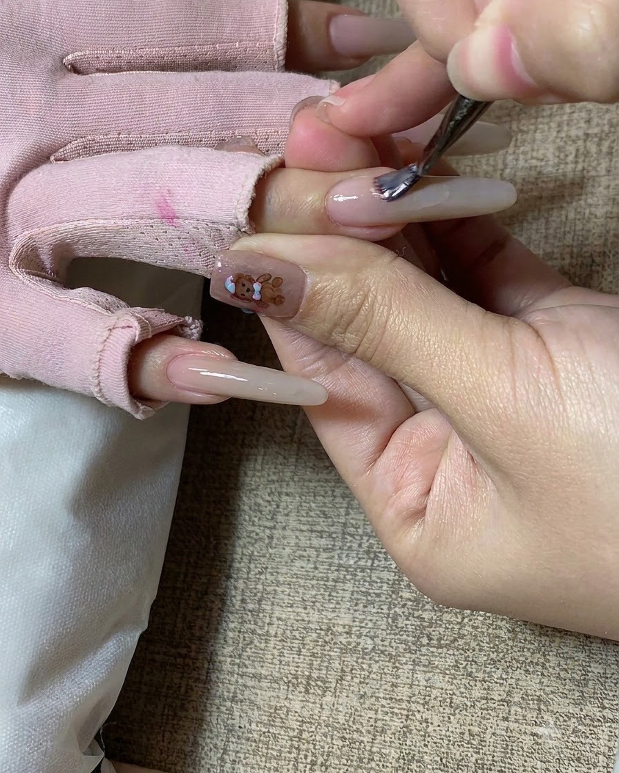 A nail brush applying milk tea colored gel onto a long sculpted fingernail.