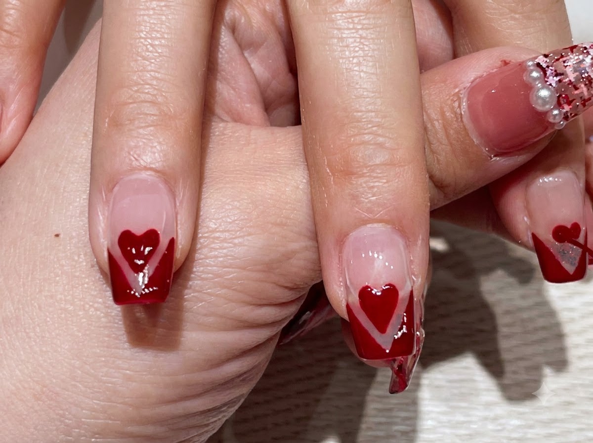 A close-up view of a hand displaying fingers with red V-tip French manicures and centered red hearts.