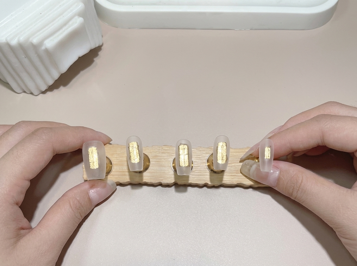 A close-up of a brush applying base gel to multiple nail tips mounted on a wooden stand.