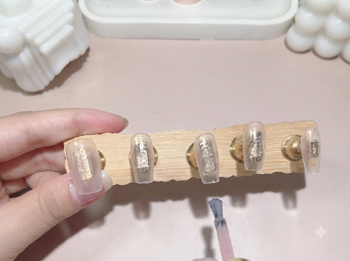 A gel polish brush applying a second coat of milky translucent pink color to nail tips attached to a wooden display stand.