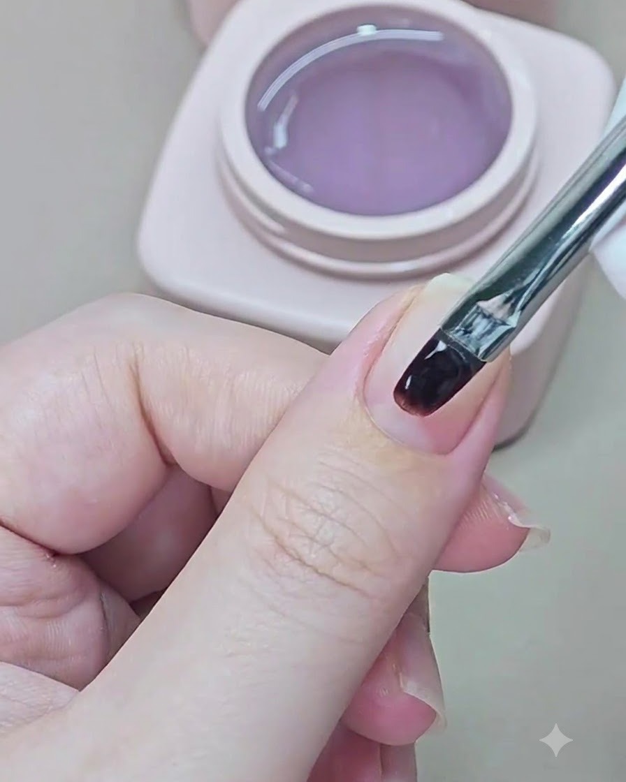 A small nail brush applying clear base gel onto a natural fingernail.
