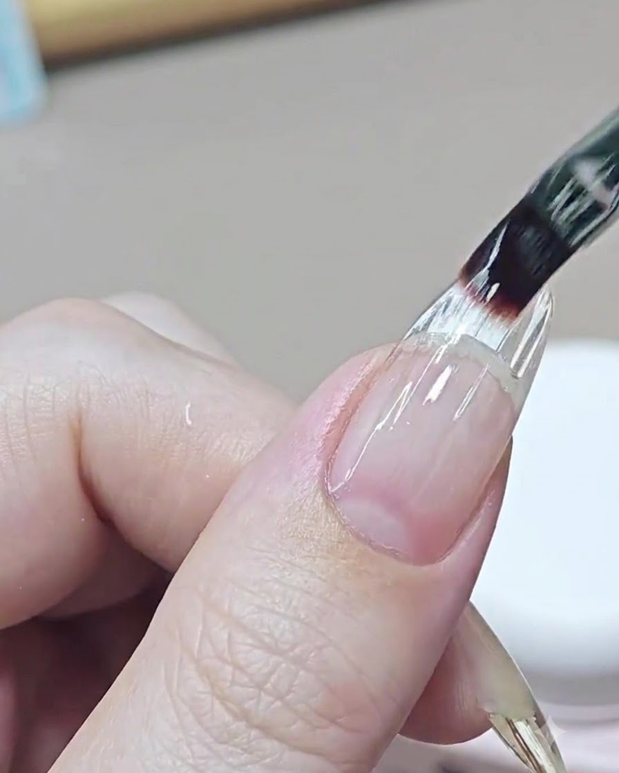 A fine nail brush applying a clear leveling layer of builder gel over the transition area of an extended nail.