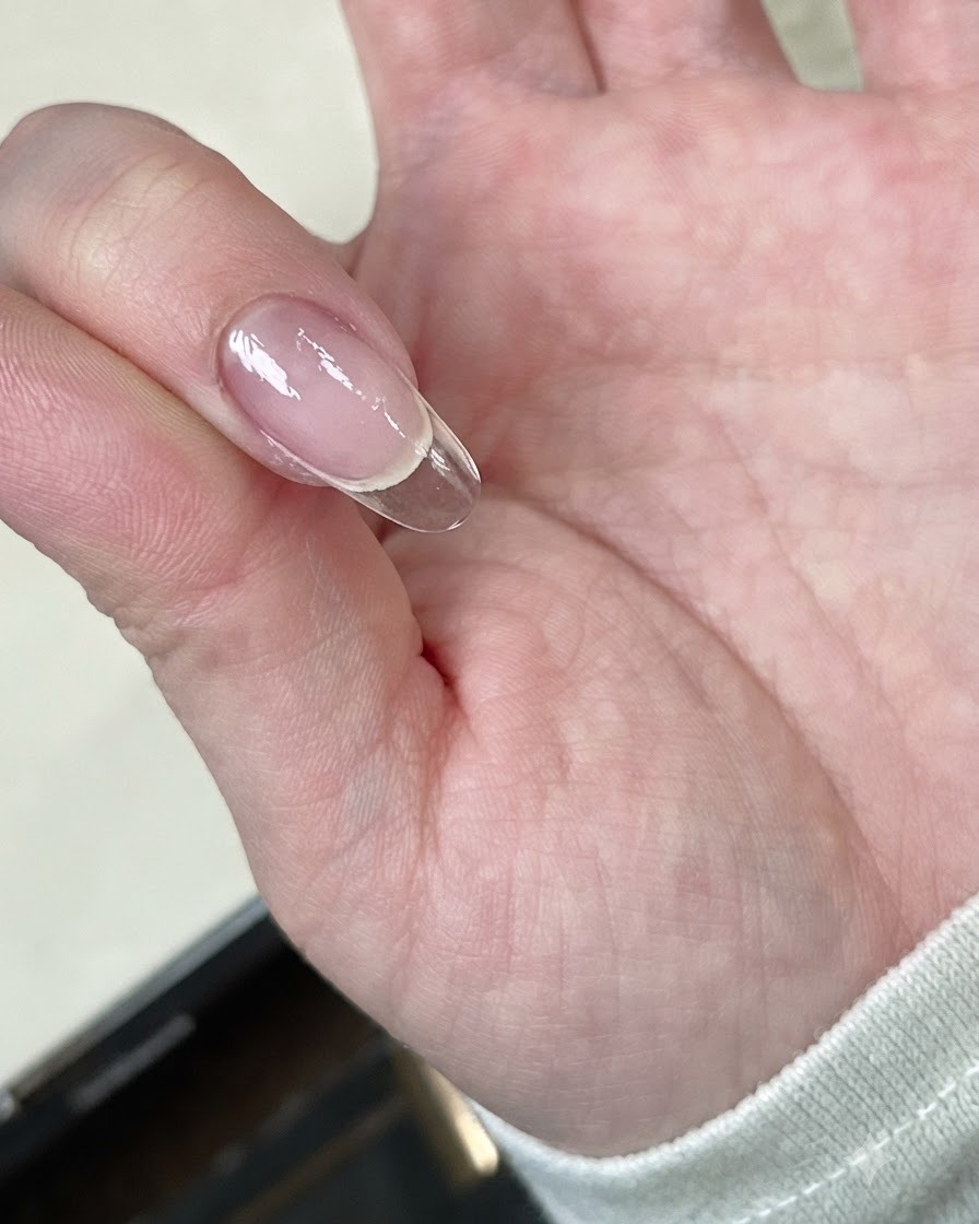 A hand holding a clear nail tip under a mini LED curing lamp.