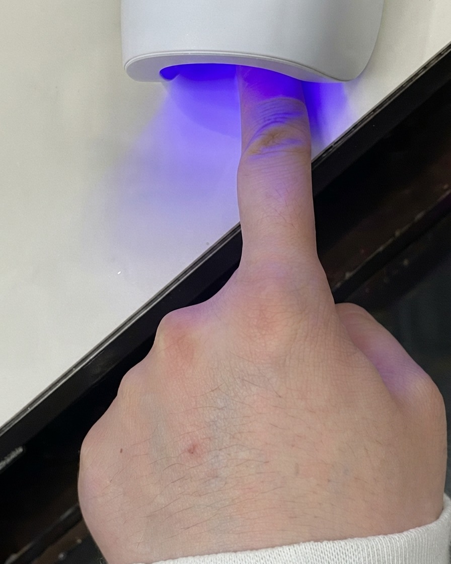 A finger placed under a mini UV LED lamp curing the gel layer.