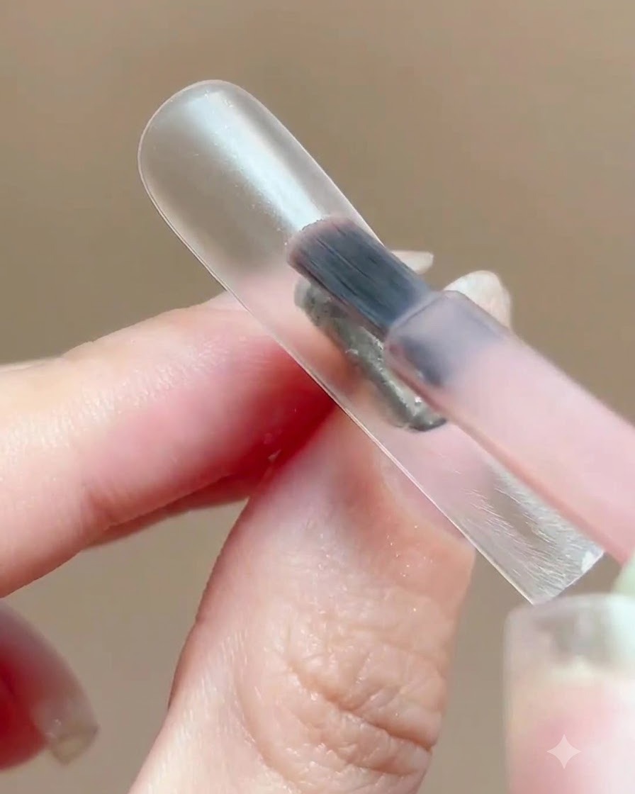 A brush applying a thin layer of translucent pink gel polish onto a clear nail display stand.