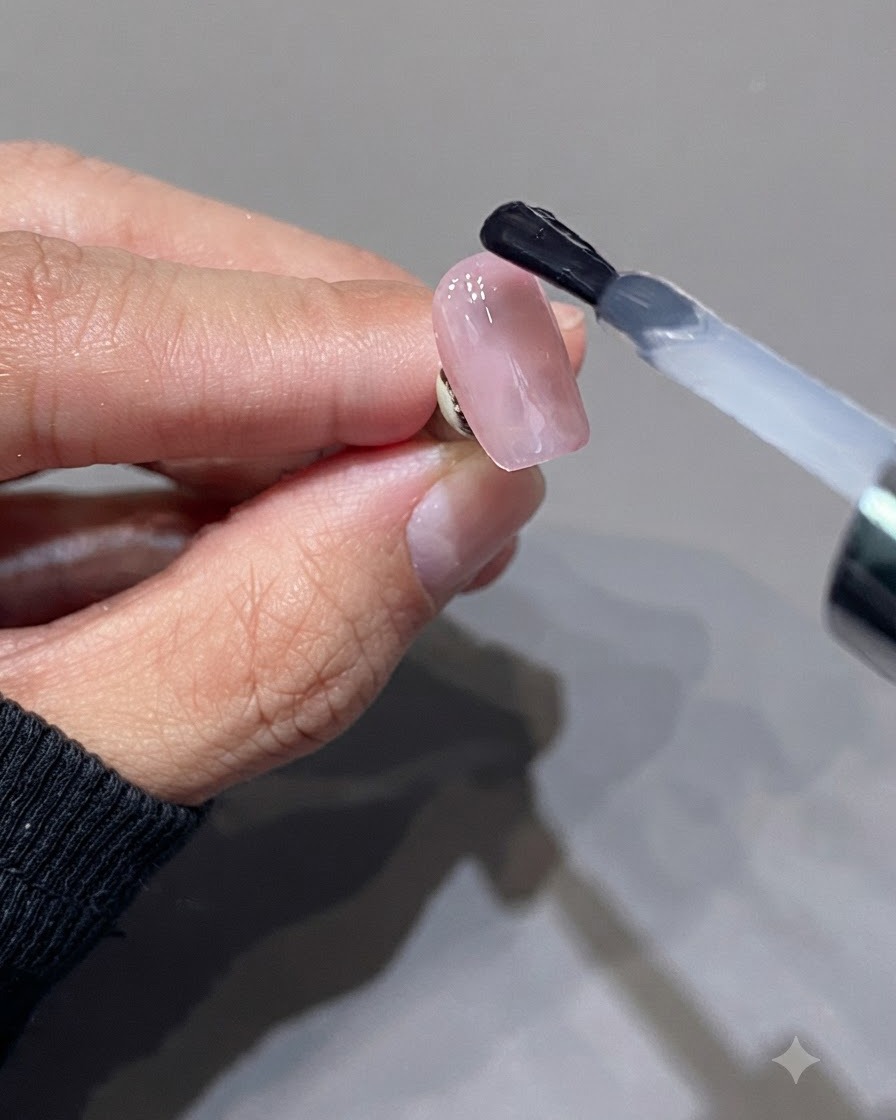 A brush applying a translucent milky pink top coat over a glossy pink nail surface.