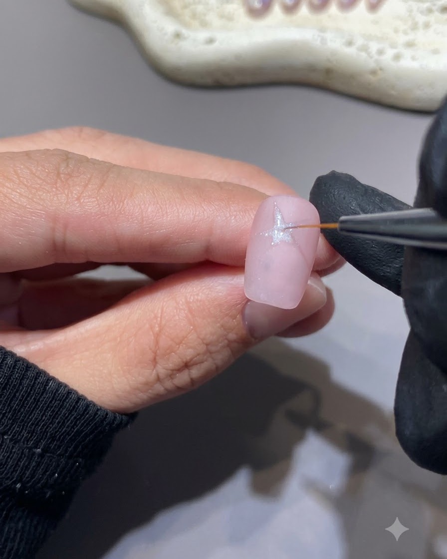 A fine liner brush drawing a white star shape onto a pink nail surface.