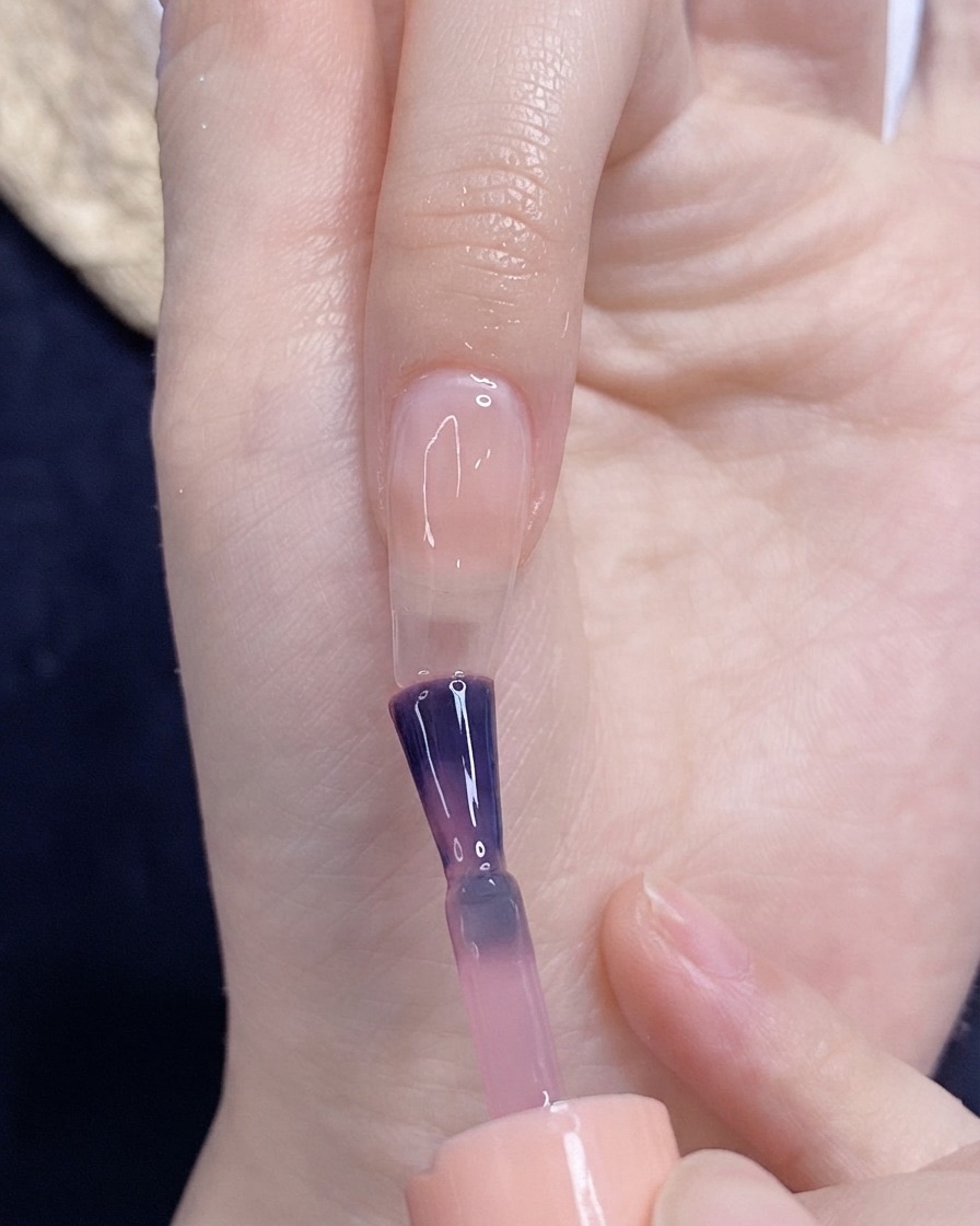 A brush applying translucent pink gel polish onto a bare fingernail.