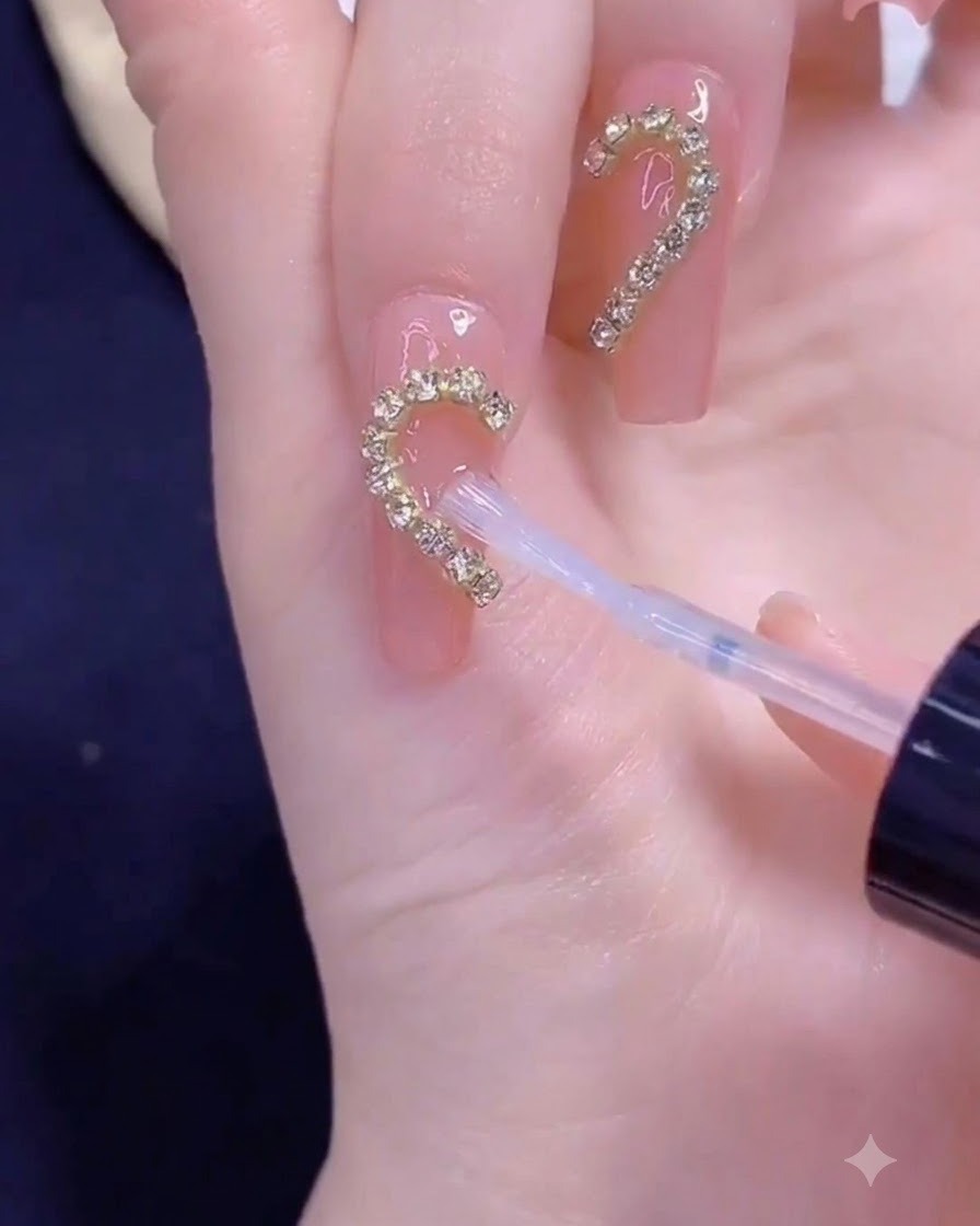 A brush applying clear top coat over a golden chain strip arranged in a half heart shape on a fingernail.