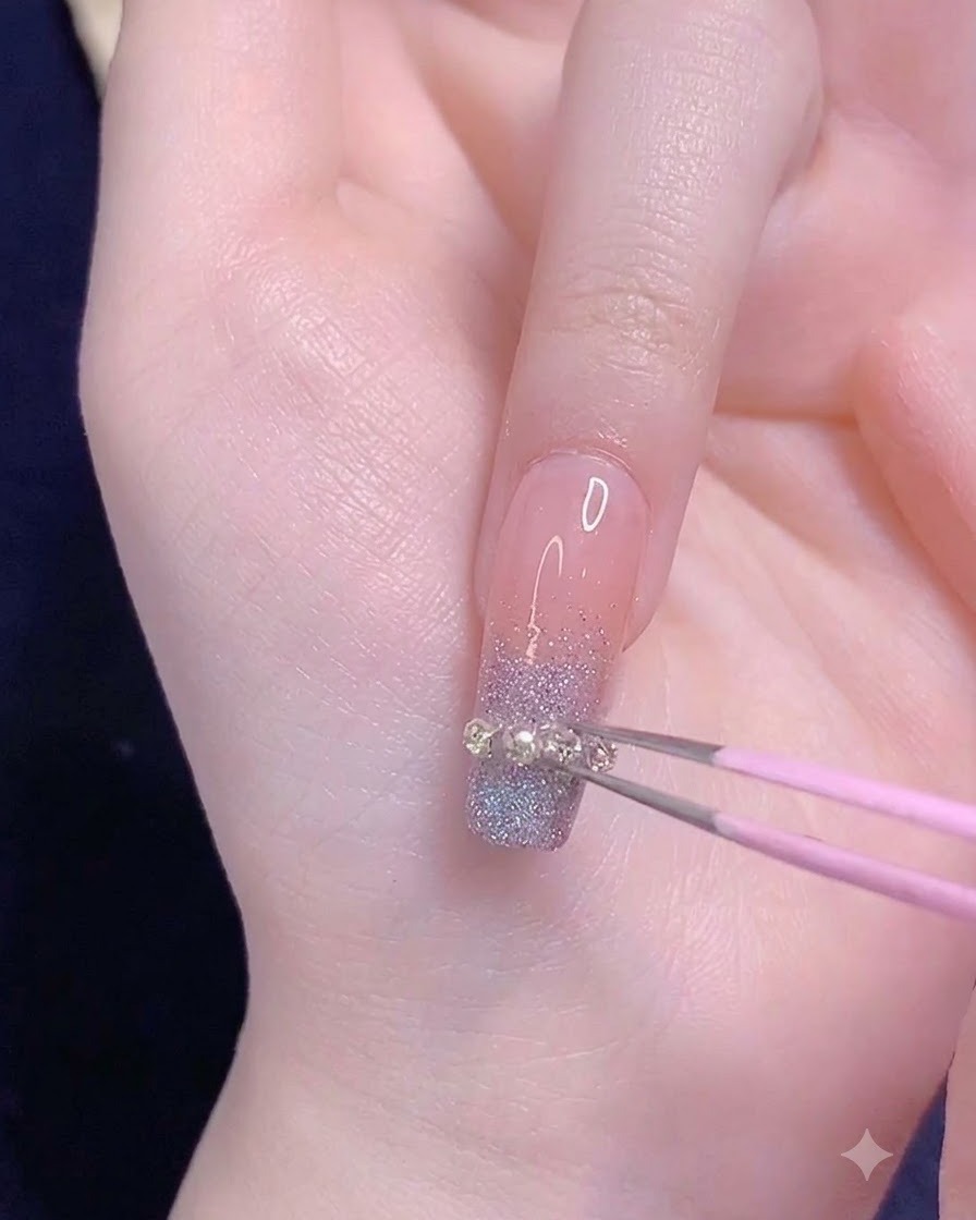 A pair of pink tweezers placing a small metallic chain strip horizontally across the gradient tip of a ring fingernail.