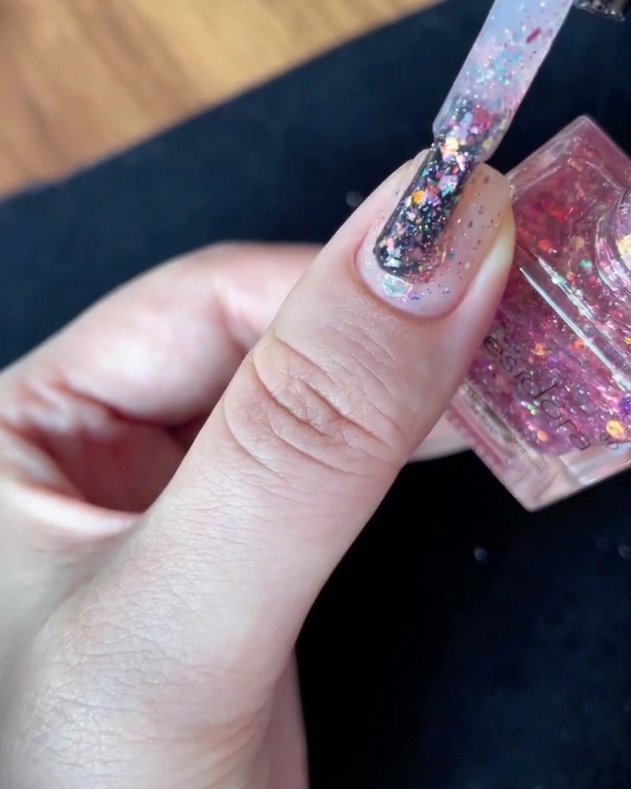A polish brush filled with glitter flakes held steady just above the surface of a nude-based nail.