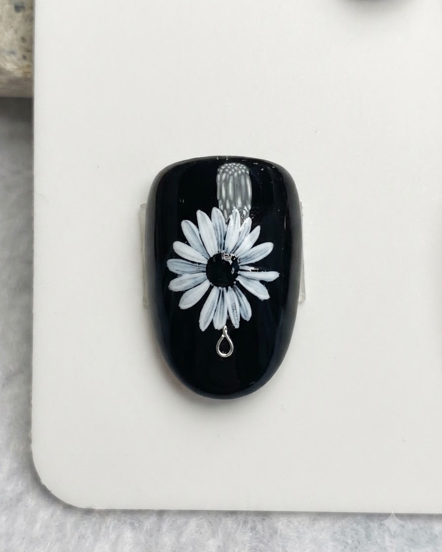 A close-up of a nail brush painting delicate white and yellow dots onto the black center of a white daisy nail art design.