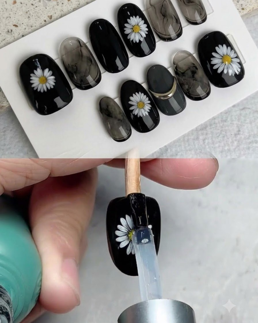 A brush applying a glossy top coat over a finished black press-on nail decorated with a white daisy.