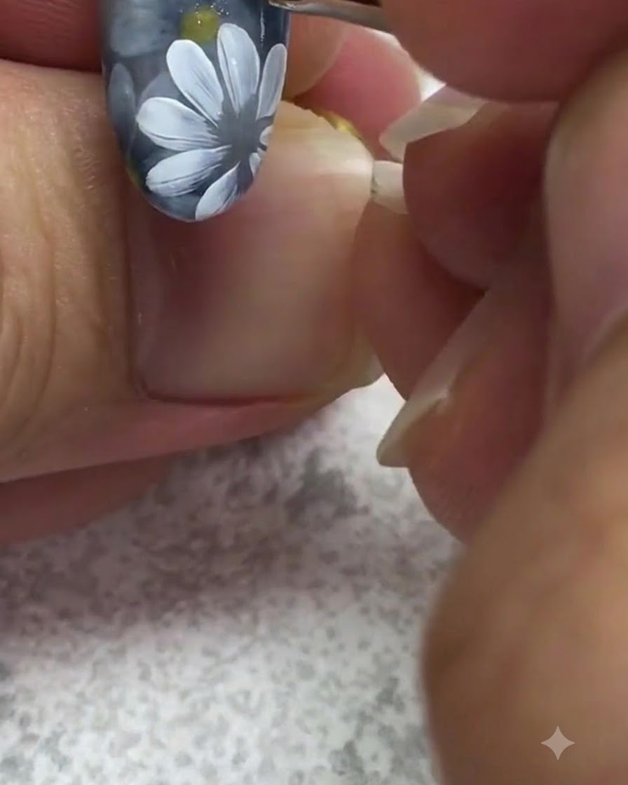 A fine detail brush applying a bright yellow dot of gel to the center of a painted white daisy on a grey nail.