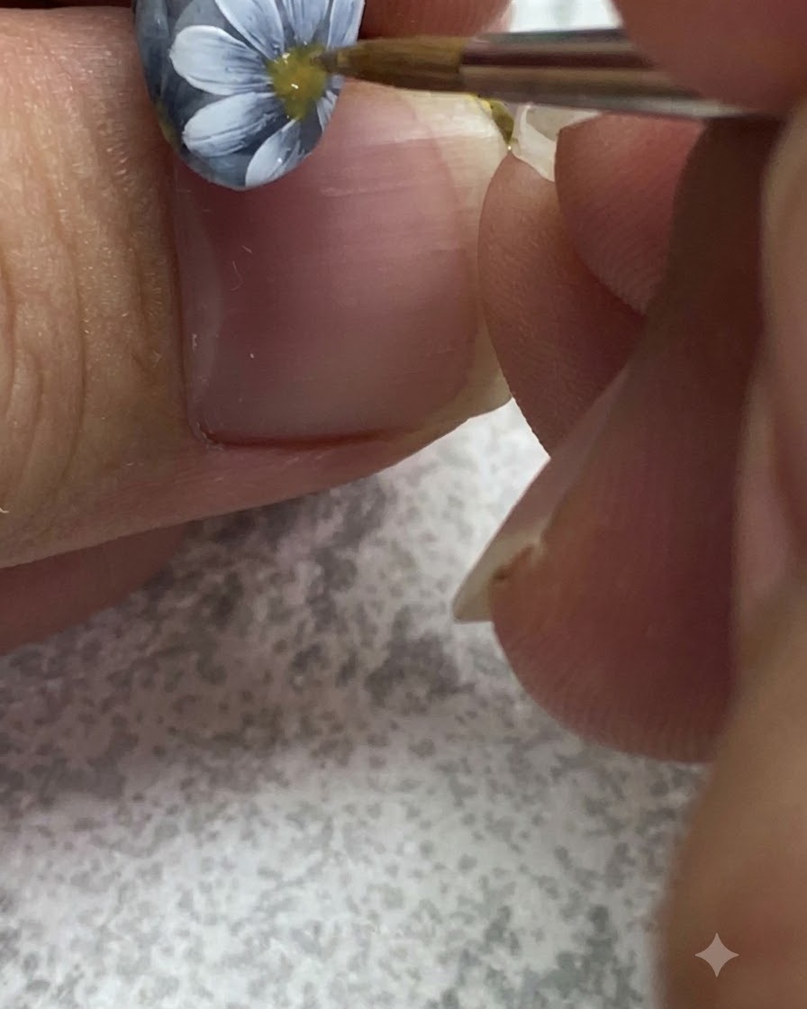 A fine brush adding detailed texture and dots inside the yellow center of a painted daisy on a grey nail.