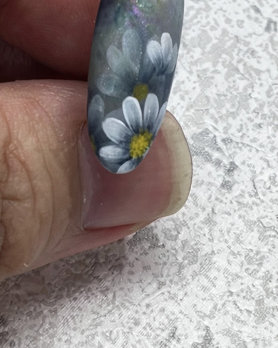 A close-up of a nail brush pulling outward to create a sharp leaf tip next to painted Nordic style daisies.