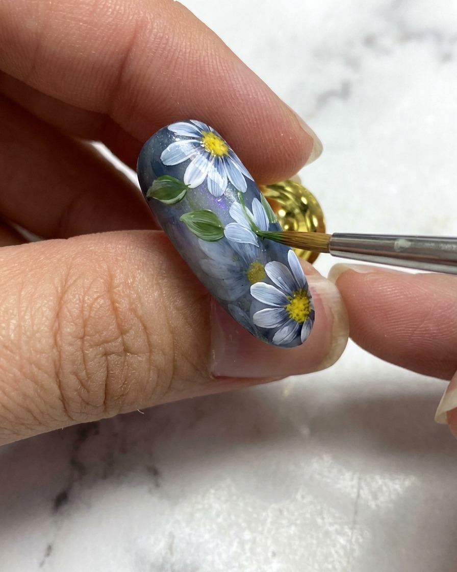 A nail art brush adding a small green leaf next to white floral details on a grey nail base.