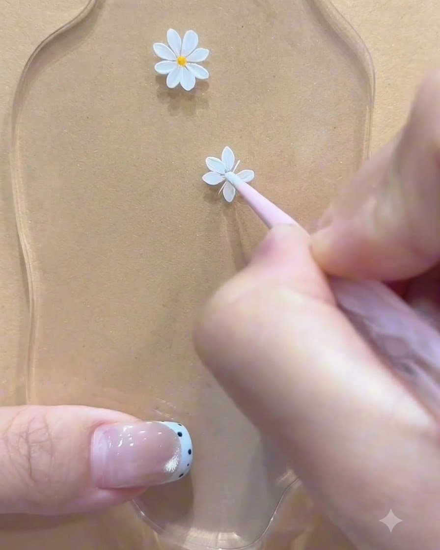 A dotting tool applying a yellow center dot to the first painted white daisy on a palette surface.