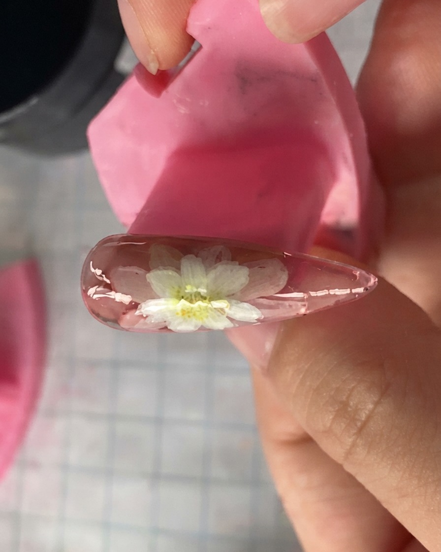 A close-up view of a pink nail model showing layered white daisy petals painted on top.