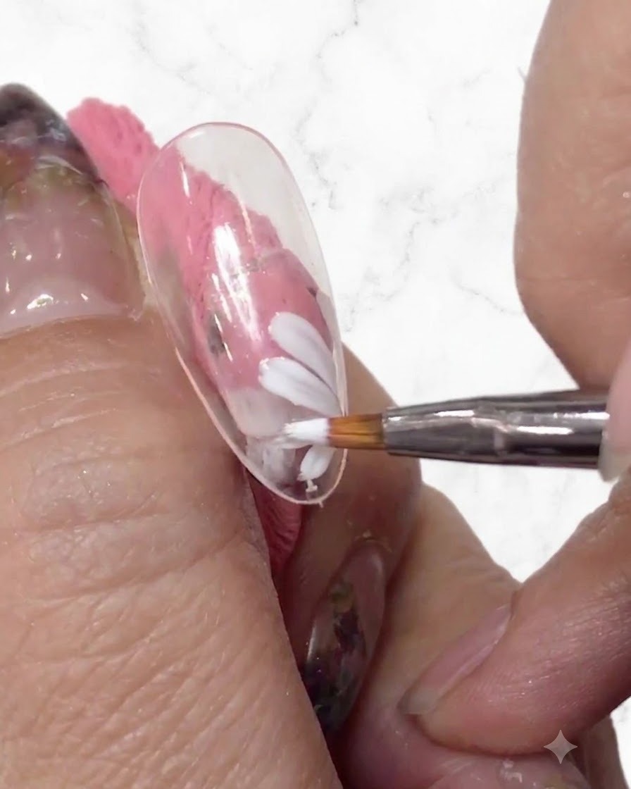 A close-up of a nail artist adding more white petals to the daisy design with a fine brush on a clear nail tip.