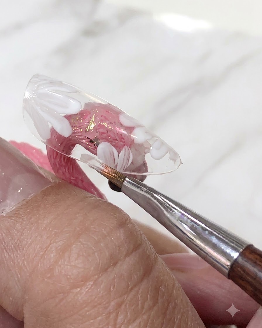 A brush applying a translucent pink gel polish layer over the painted white flowers on a clear nail tip.