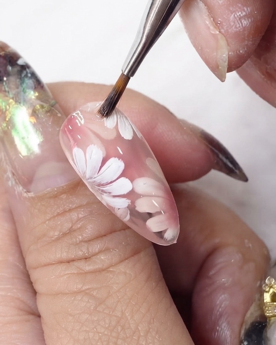 A gel brush gliding a coat of sheer pink polish over painted white flowers on an almond-shaped nail.
