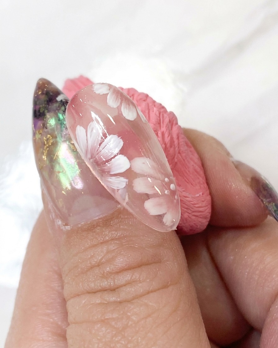 A dotting tool applying tiny white accent dots around the flower center of a detailed nail art design.