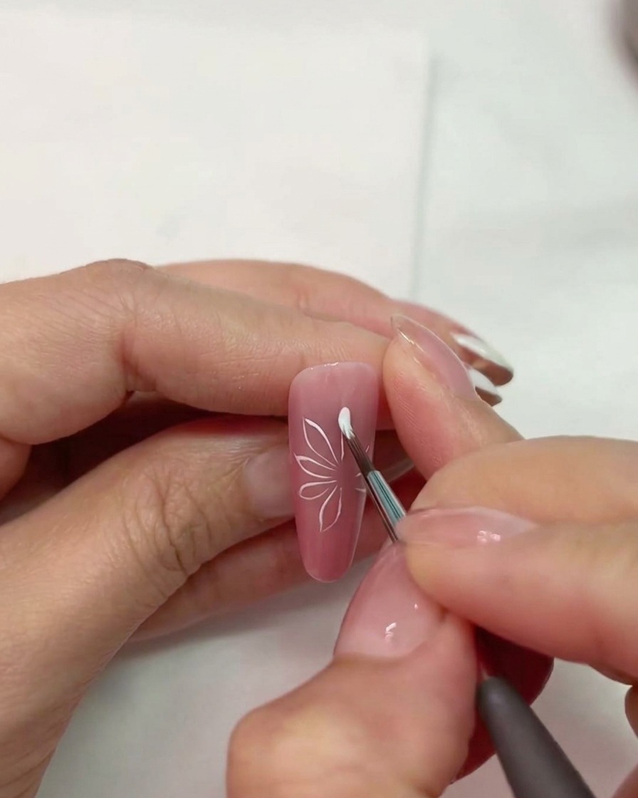 A close-up of a nail brush drawing white petal shapes on a pink matte nail.