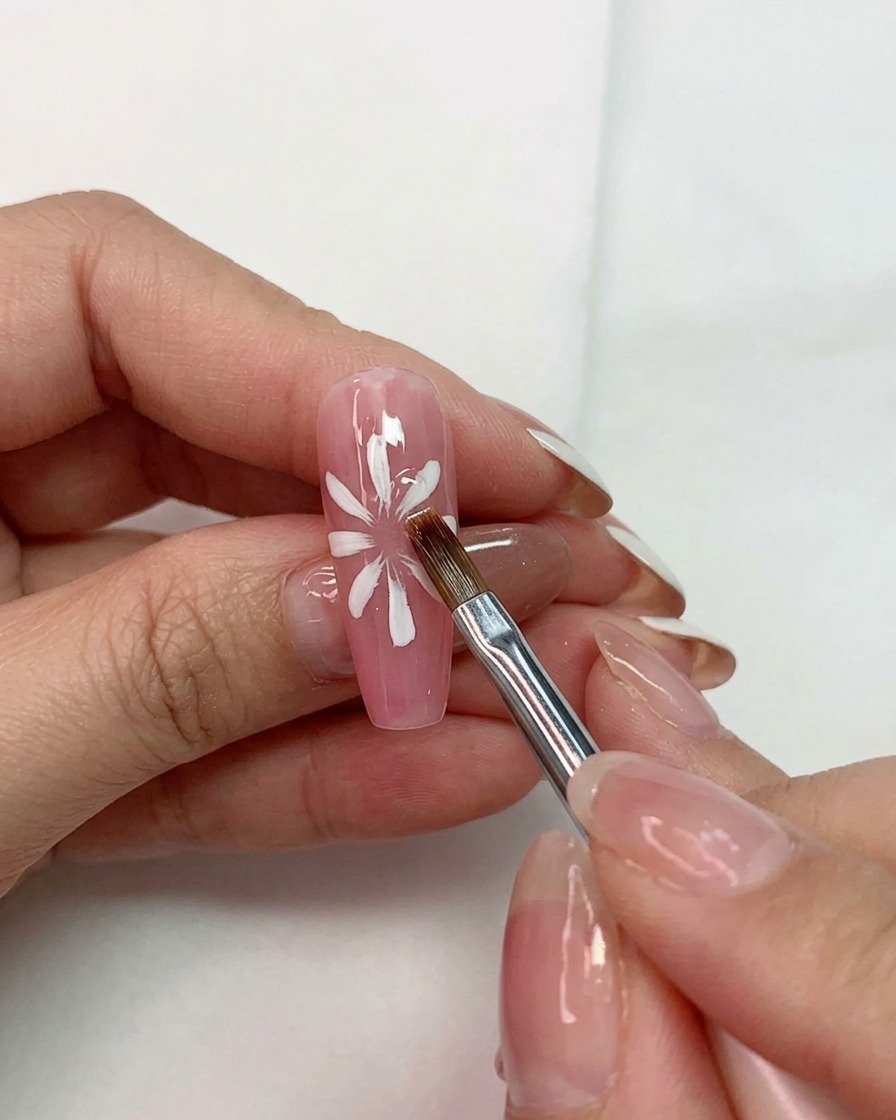 A hand holding a multi-purpose brush shaping white petals over a pink base on a fingernail.