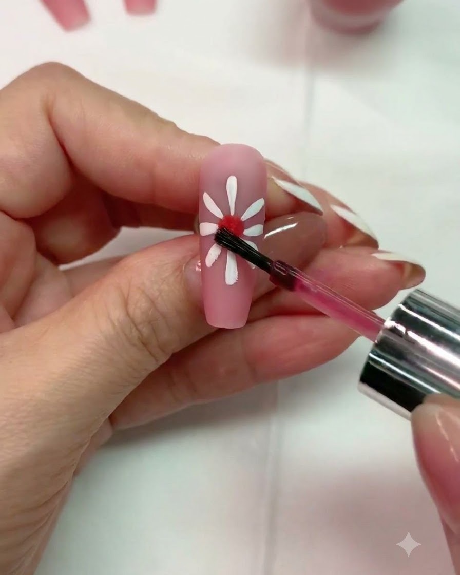 A brush applying a small drop of red blooming gel to the center of a white daisy design on a matte pink nail.
