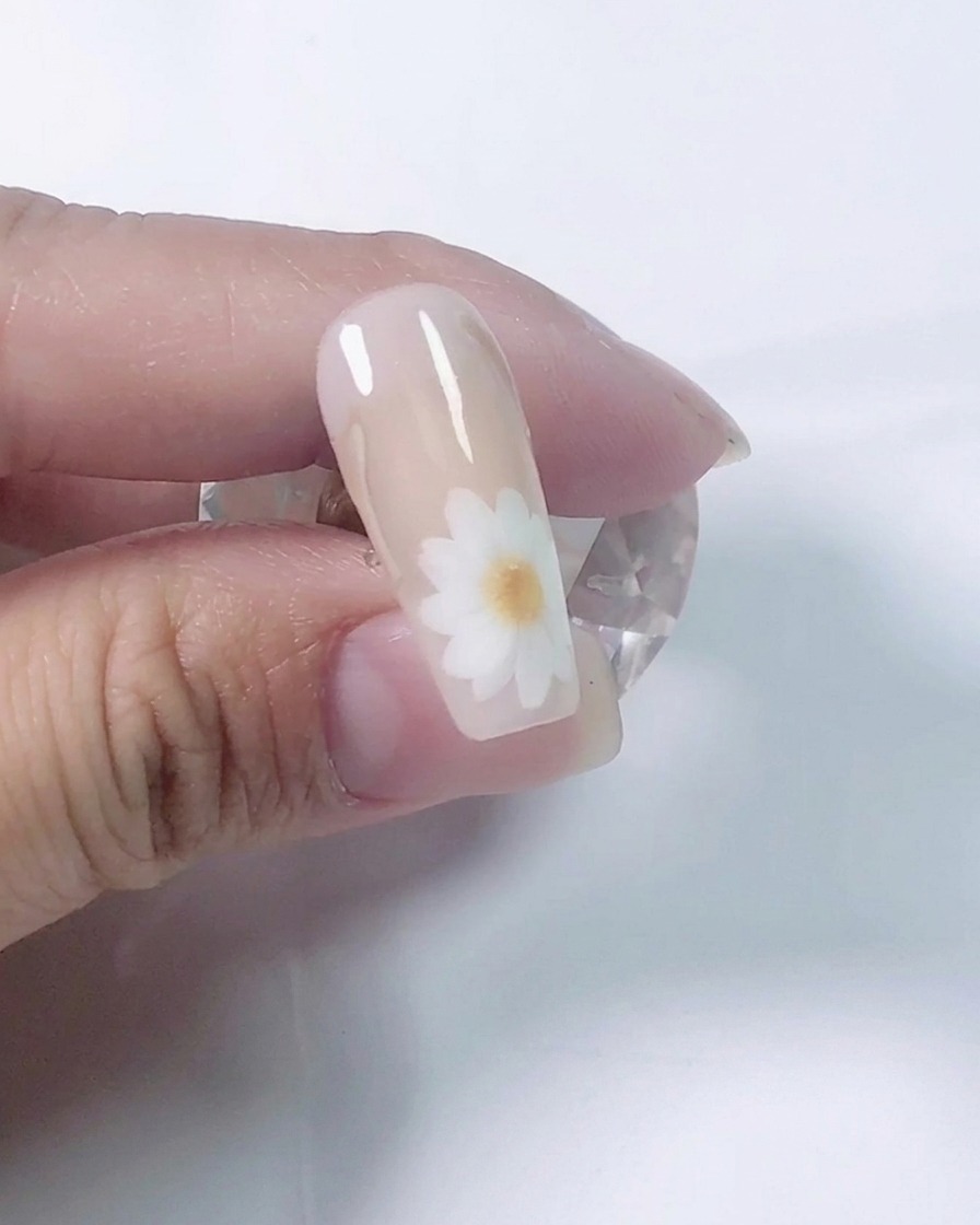 A close-up shot of a nail artist using a fine brush to paint white daisy petals on a nude practice nail.